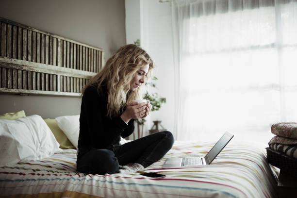 frau mit laptop-computer sitzt auf dem bett zu hause - websurfen stock-fotos und bilder