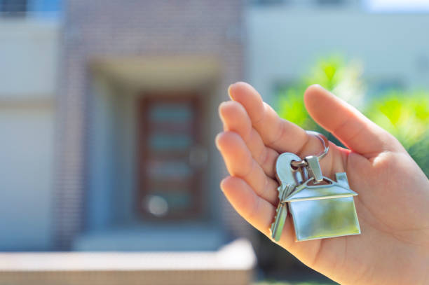 Hand holding a house key in front of a large house.