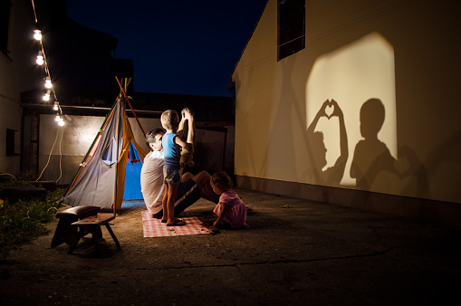 Father,brother and sister are playing with shadows in backyard of their home. There is tent, lights and good mood.