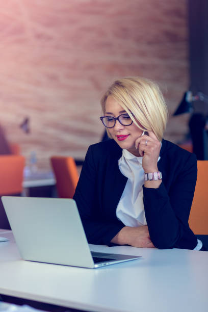 istock Attractive charming blonde business woman talking on a mobile while sitting at office desk working on a laptop. 918746536