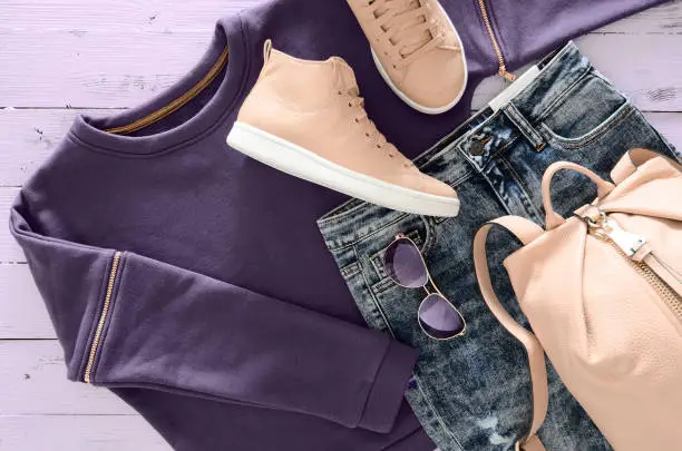 Photo of Womens clothing, accessories, footwear (violet sweatshirt, acid washed jeans,  leather backpack and sneakers, sunglasses) on wooden background. Outfit for teens. Top view, flat lay. Trendy colors