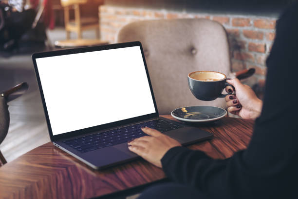 Mockup image of a businesswoman using laptop with blank white desktop screen while drinking hot coffee on wooden table in cafe
