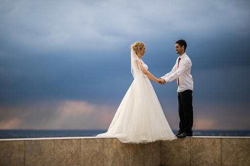 Young happy newlyweds standing on the edge of a terrace while holding hands and communicating. Copy space.