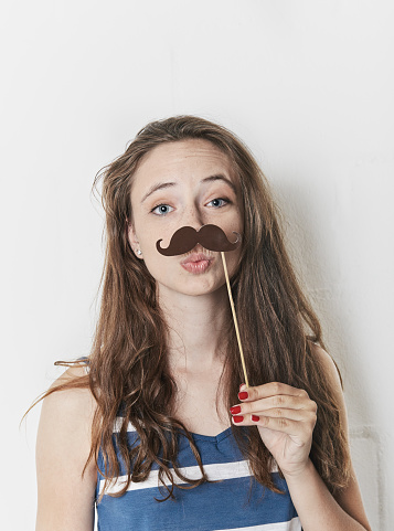 A cute young woman pulls a funny face as she holds a fake moustache under her nose.