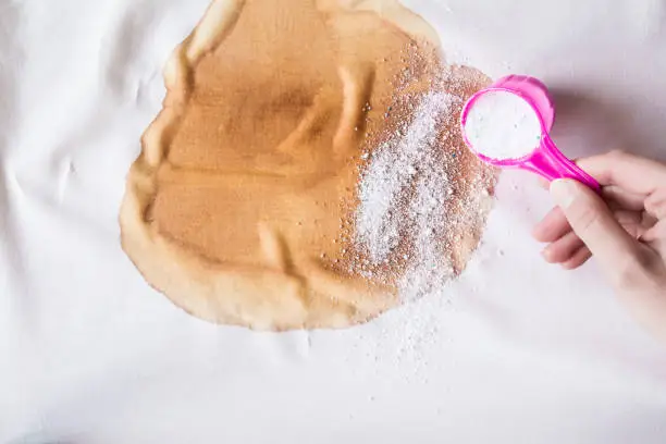 Photo of Big, brown coffee stain on light, white cloth. Woman's hand strewing washing powder from a cup or scoop. Spilled beverage stain removing. Dry cleaning concept. Clothes care. Chores of housewife.