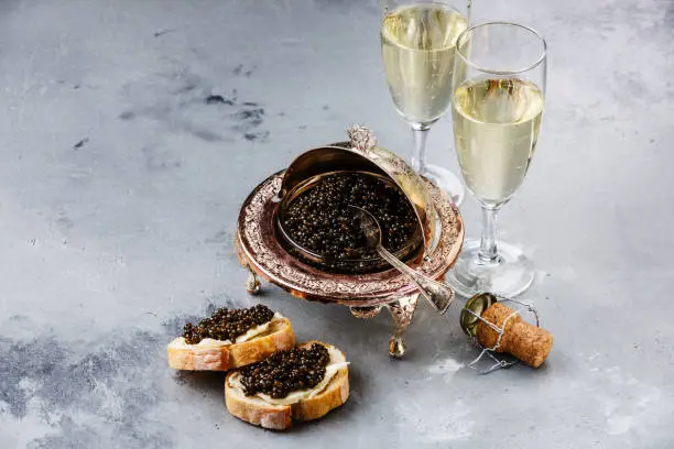 Black caviar in silver bowl, sandwiches and champagne on gray concrete background