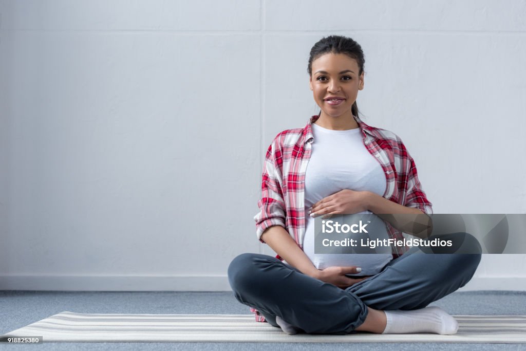 americana africano mujer embarazada sentada en la estera de la yoga - Foto de stock de Embarazada libre de derechos