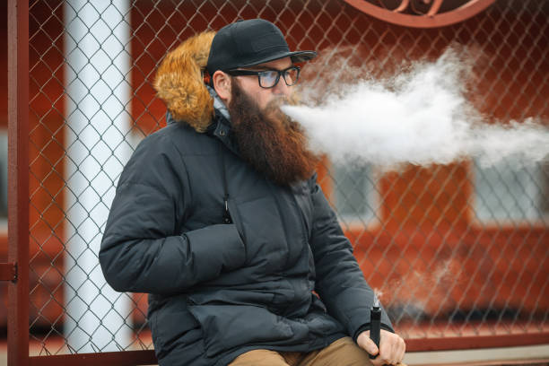 istock Young guy with large beard in glasses and a cap vaping an electronic cigarette. 918855344