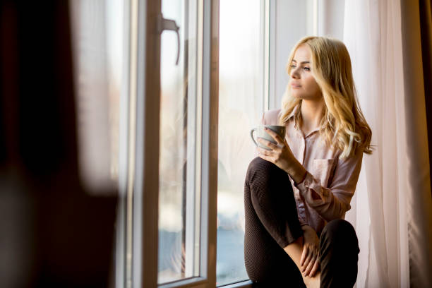 istock Blonde young woman drinking coffee by window 918881044