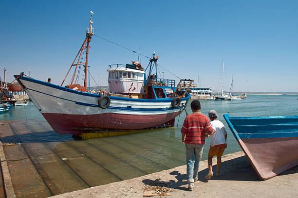 Arrecife, Lanzarote, Canary Islands, Spain - February 28th 2025:   A vibrant fishing vessel rests in the bustling harbor of Arrecife, its red, white, and blue hues standing out against the calm waters. Equipped with essential fishing gear, the boat reflects the maritime traditions of the Canary Islands. In the background, a mix of smaller boats and a towering cruise ship highlight the diversity of vessels navigating the port. The partly cloudy sky adds depth to the scene, capturing the essence of coastal life in this picturesque harbor.