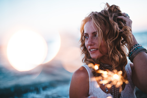Hippie woman with jewelry and wavy hair celebrating with sparkers at beach festival at sunset