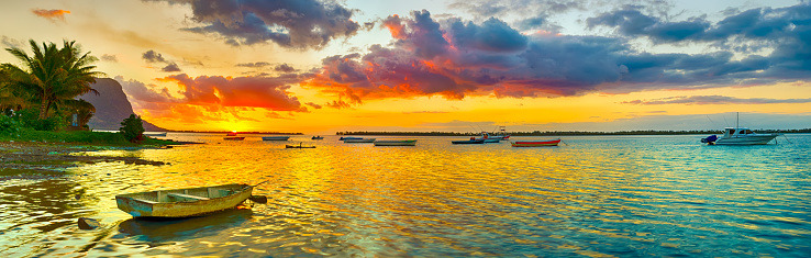 A panoramic view of a tropical ocean with boats scattered throughout. The ocean is a beautiful turquoise color and the sky is blue with white fluffy clouds. The boats are different colors and sizes, some are closer to the shore and some are further out. The shore is sandy and there are trees and buildings in the background. The photo is taken from a high vantage point and the horizon is visible.