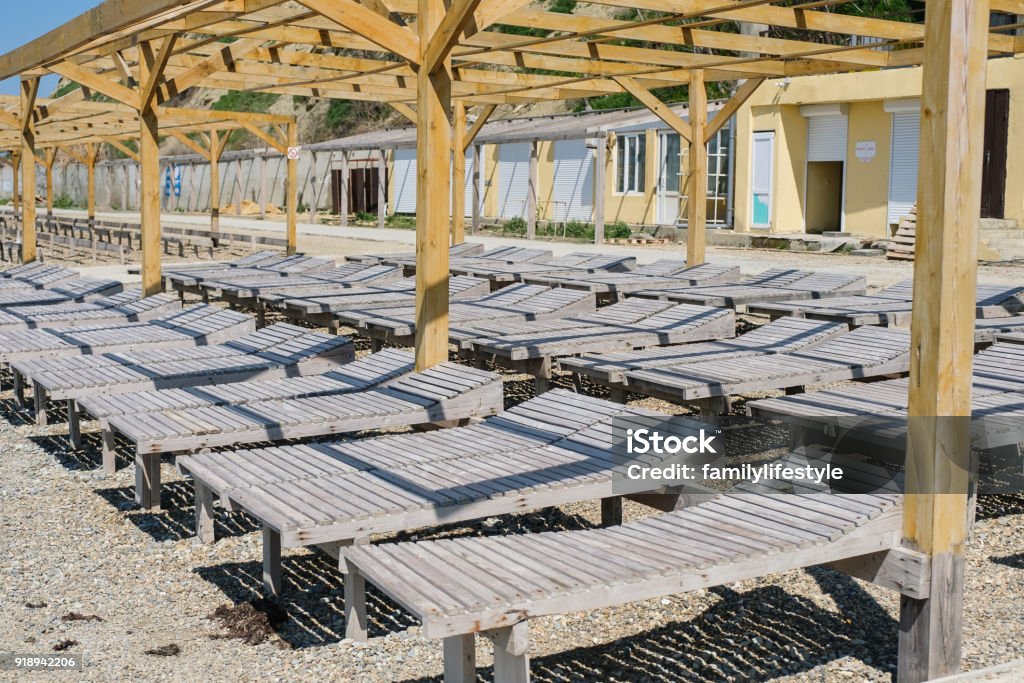 Wooden sun loungers on a pebbled public beach by the sea. Close-up side view. Wooden sun loungers on a pebbled public beach by the sea. Side view. Beach Stock Photo