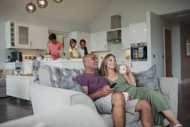 Husband and wife relaxing on the sofa while the children prepare food in the kitchen.