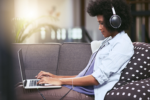 Shot of a young woman using a laptop and headphones on the sofa at home