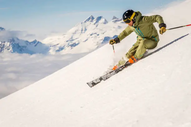 A skier carving down a ski run in the european alps, snow capped mountains and cloud covered valleys in the background.