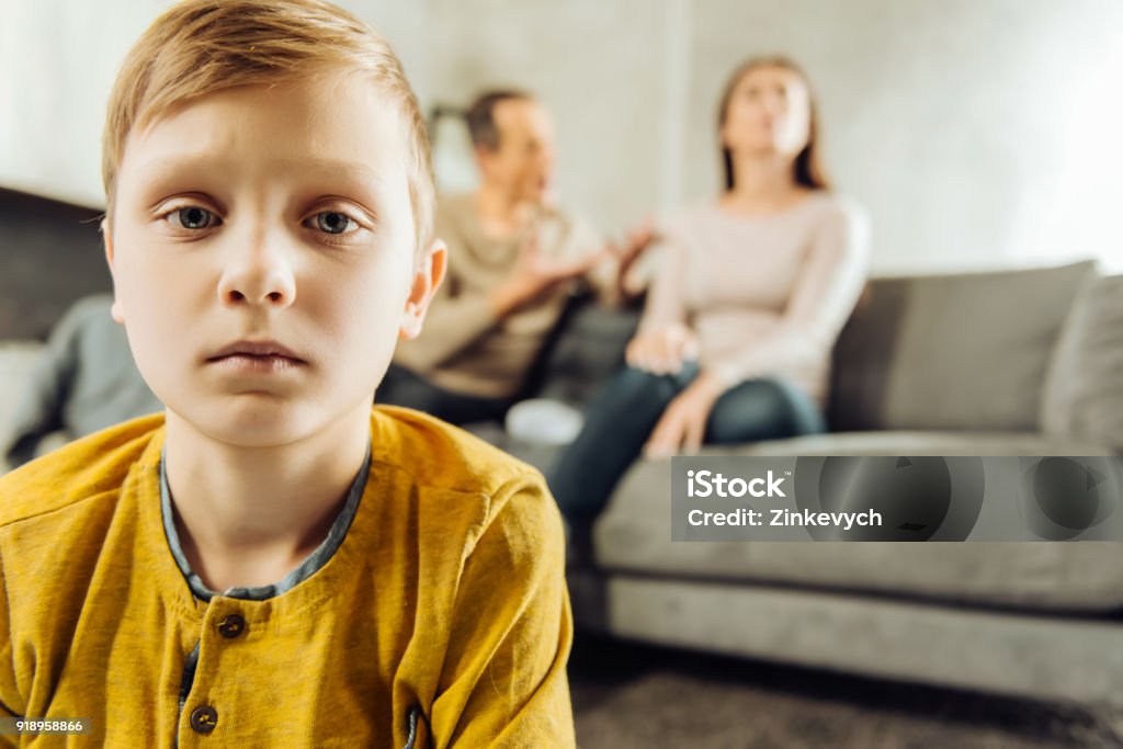 Little boy feeling lonely while his parents quarrelling All alone. The focus being on the upset little boy feeling lonely and looking sadly at the camera while his parents sitting on the couch and quarrelling in the background Child Stock Photo