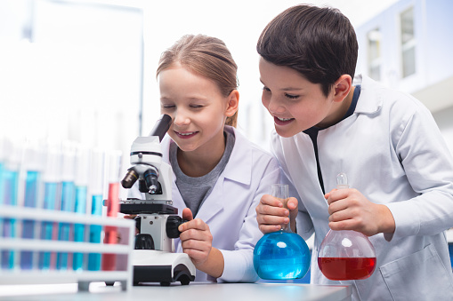 Precise look. Jolly energetic curious girl using microscope and smiling while boy holding bulbs