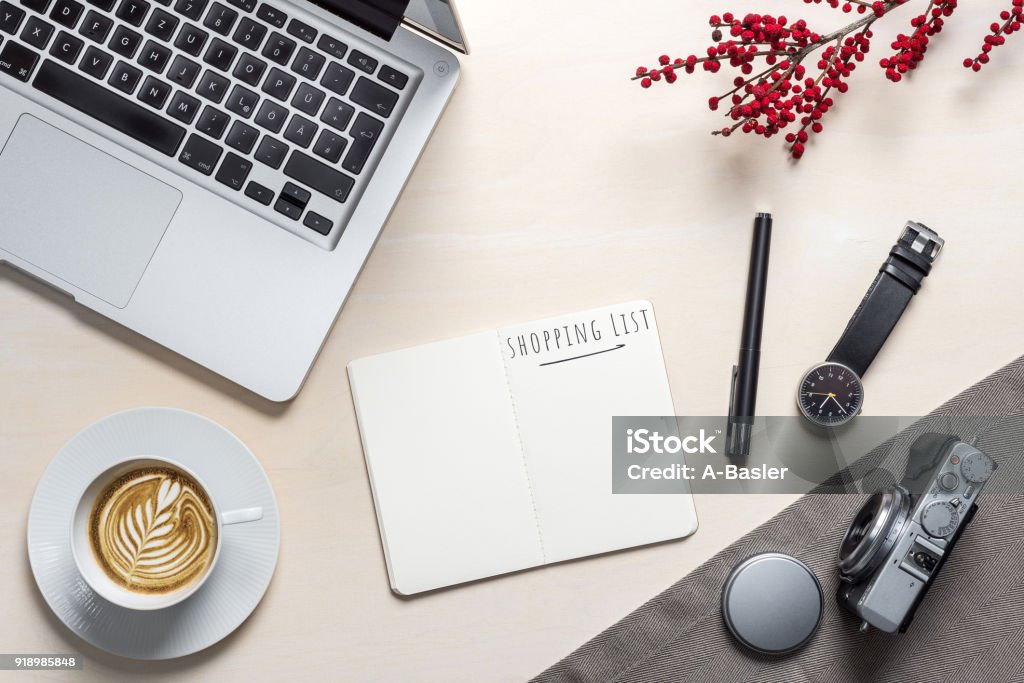 Shopping list written on open notepad in office as flatlay Shopping list writing in open notepad with coffee mug, camera, clock and laptop lying on photographer office desk as flat lay Above Stock Photo