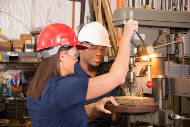 African descent and Latin descent male and female factory workers use a industrial drill press machine in a small factory setting.  The mid-adult team of machinists wear blue uniforms, hard hats, safety glasses.
