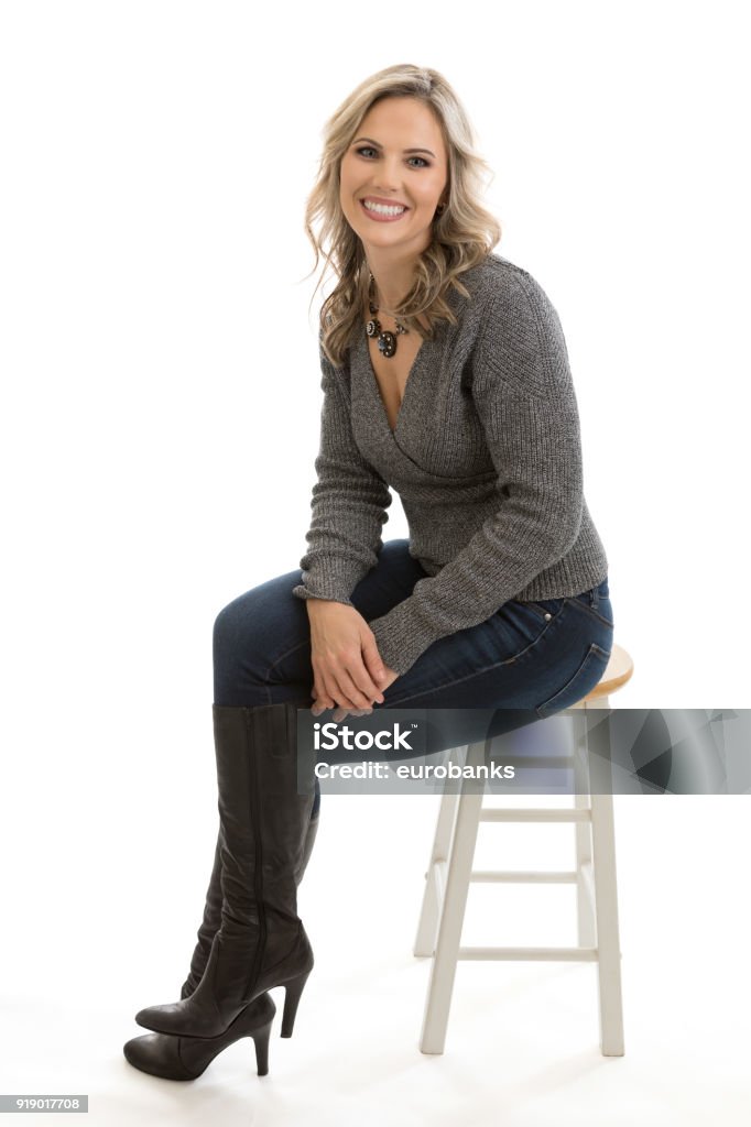 Beautiful Woman Sitting on a Stool Full length portrait of a beautiful mid 30s blond woman sitting on a stool wearing sweater and blue jeans isolated on white background Women Stock Photo