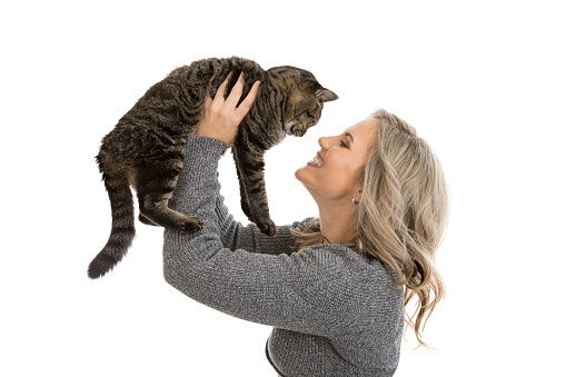 Beautiful blond mid 30s woman holding her adorable grey tabby cat in the air, nose to nose, isolated on white background