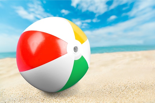 Stock photo showing close-up view of sandy beach with a multicoloured, rainbow striped football by water's edge of sea at low tide in the background. LGBTQ+ gay holiday concept.