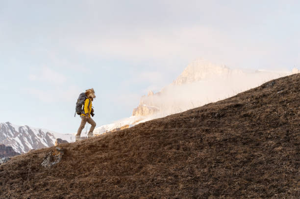 istock A backpacker in a big fur hat and gloves with a backpack on his back goes uphill against the background of epic cliffs in the clouds. The concept of mountain tourism in any season 919306988