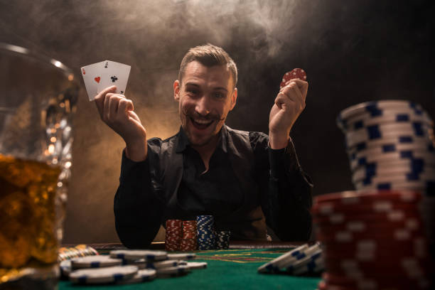handsome poker player with two aces in his hands and chips sitting at poker table in a dark room full of cigarette smoke - smiling casino human hand beautiful imagens e fotografias de stock