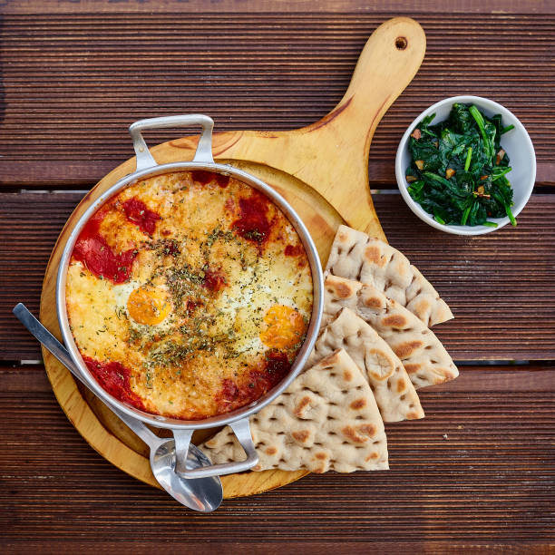 Scrambled eggs with tomatoes, cheese and spices in an aluminum frying pan with handles with homemade thin lavash and juicy greens on a wooden brown table. The photo was taken under natural light.