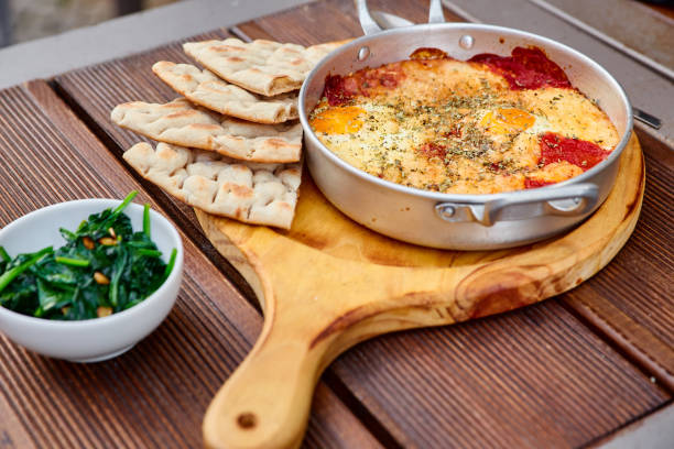 Scrambled eggs with tomatoes, cheese and spices in an aluminum frying pan with handles with homemade thin lavash and juicy greens on a wooden brown table. The photo was taken under natural light.