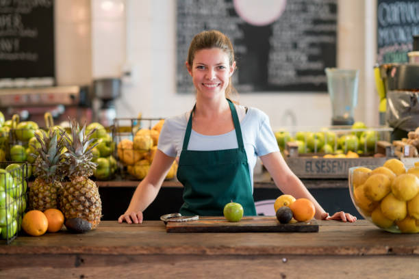 bellissimo dipendente di una juice bar in piedi dietro il bancone con mele, lime, arance, ananas sul tavolo guardando la telecamera sorridente - orange uniform foto e immagini stock