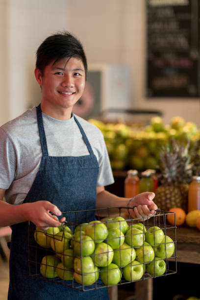 istock Asian employee at a juice bar organizing the fruit baskets looking at camera smiling while holding a basket full of apples 919359200