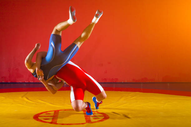Two strong wrestlers in blue and red wrestling tights are wrestlng and making a suplex wrestling on a yellow wrestling carpet in the gym. Young man doing grapple.