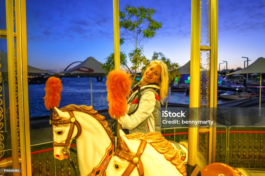 Carousel in Elizabeth Quay Happy blonde caucasian woman riding on white horse of carousel, at Esplanade in Perth, Western Australia. Elizabeth Quay by night. Leisure and recreational concept. Adult Stock Photo