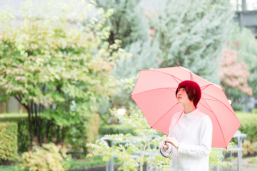 Asian woman with a red umbrella is walking in the garden of the office building. She wears casual white collar shirt.