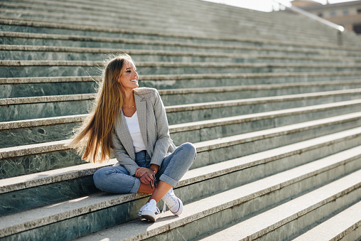 Beautiful young caucasian woman smiling in urban background. Blond girl wearing casual clothes in the street. Female with elegant jacket and blue jeans sitting on stairs.