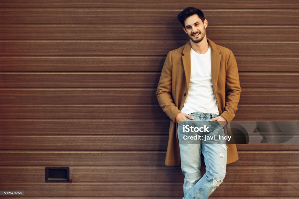 Smiling man wearing demi-season clothes in the street. Young smiling man wearing demi-season clothes in the street. Young bearded guy with modern hairstyle with coat, blue jeans and t-shirt against urban wooden blinds. Men Stock Photo