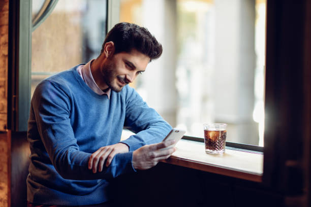 istock Smiling man with blue sweater looking at his smartphone in a modern pub. 919583342