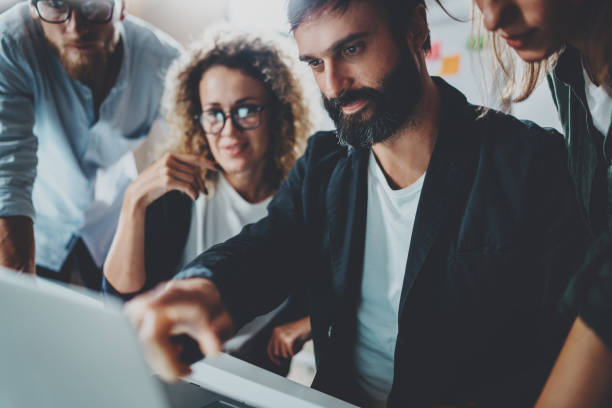 istock Coworkers working together in modern office.Bearded man talking with colleague and using mobile laptop while sitting at the wooden table.Horizontal.Blurred background.Cropped. 919641894