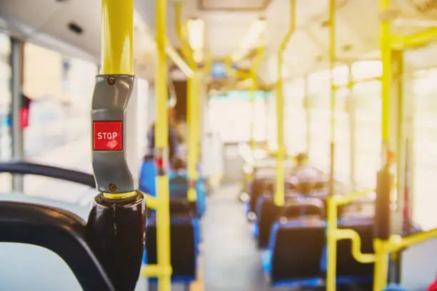 Photo of Red button STOP on the bus. Bus with yellow handrails and blue seats. Photo with the sun effect, glare on the lens from the light. Spacious interior of the bus, bright button with focus.