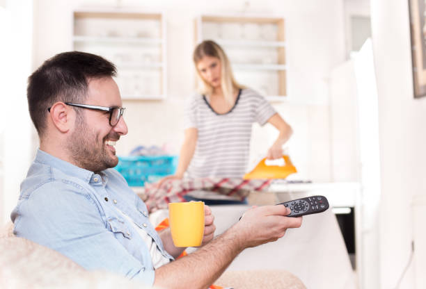 Man watching tv phone while wife doing chores Young handsome man sitting on sofa and holding tv remote control while wife ironing and doing chores in background laundry husband housework men stock pictures, royalty-free photos & images