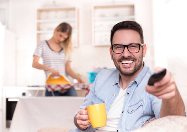 istock Man watching tv phone while wife doing chores 919694888