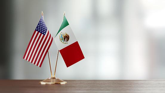 American and Mexican flag pair on desk over defocused background. Horizontal composition with copy space and selective focus.