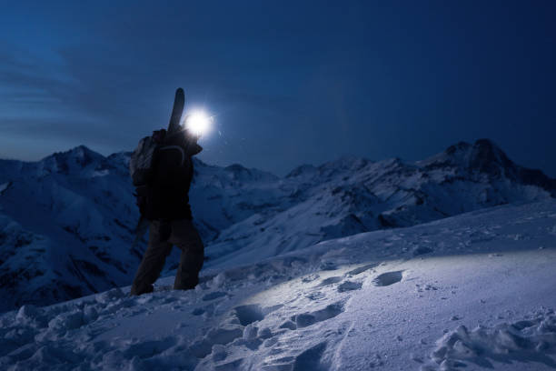 istock Professional tourist commit climb on great snowy mountain at night. Wearing backpack, headlamp and ski wear. Backcountry. Brave extreme traveler with a snowboard behind his back climbs winter slope 919758868