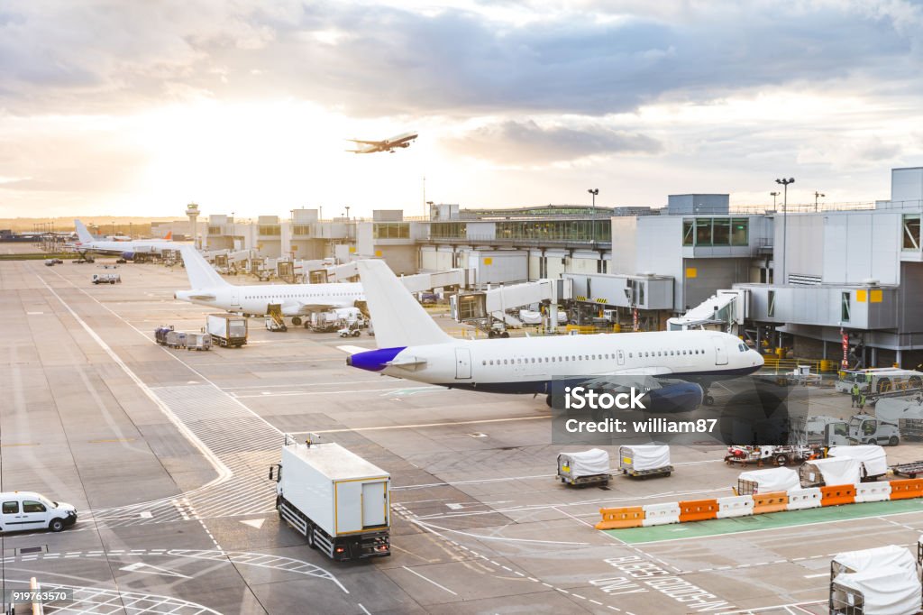 Busy airport view with airplanes and service vehicles at sunset Busy airport view with airplanes and service vehicles at sunset. London airport with aircrafts at gates and taking off, trucks all around and sun setting on background. Travel and industry concepts Airport Stock Photo