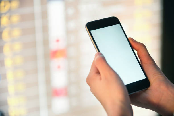 istock Mockup image of female hands holding black mobile phone with blank white screen over flight board in airport terminal 919807536