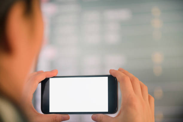 istock Mockup image of female hands holding black mobile phone with blank white screen over flight board in airport terminal 919807568
