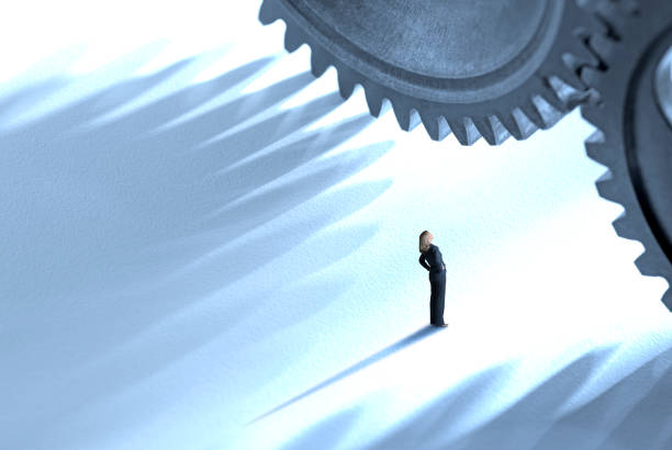 A businesswoman looks up at two large interlocking gears that dwarf her and cast their shadow across the ground around her.