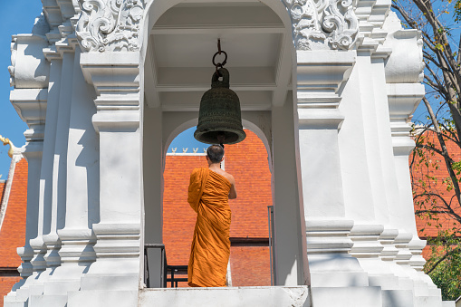 CHIANG MAI THAILAND - JANUARY 29 2018; Buddhist Monk stands  on high level of white ornate bell tower ringing bell matching orange robes and roof top behind